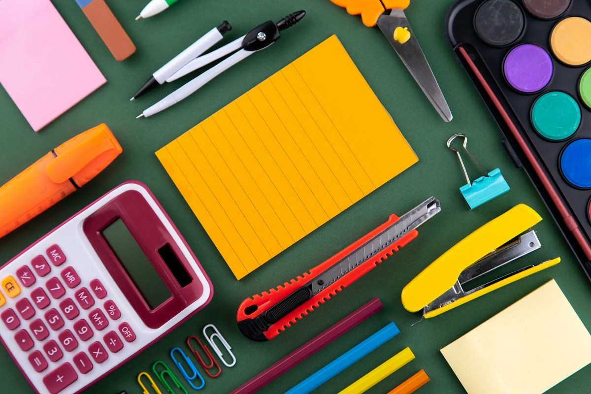 Stationery laid out on a desk: a calculator, sticky notes, pens, a stapler, scissors, a paint set and paper clips